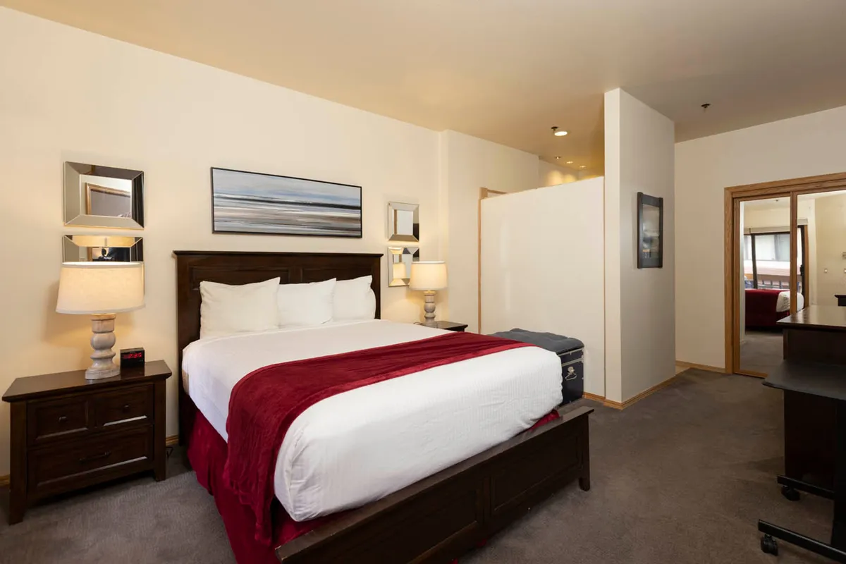 A cozy bedroom scene featuring a bed with crisp white linens and a vibrant red throw, flanked by nightstands with softly glowing lamps. Abstract artwork hangs above the bed, and natural light gently fills the room, creating an inviting and serene atmosphere.