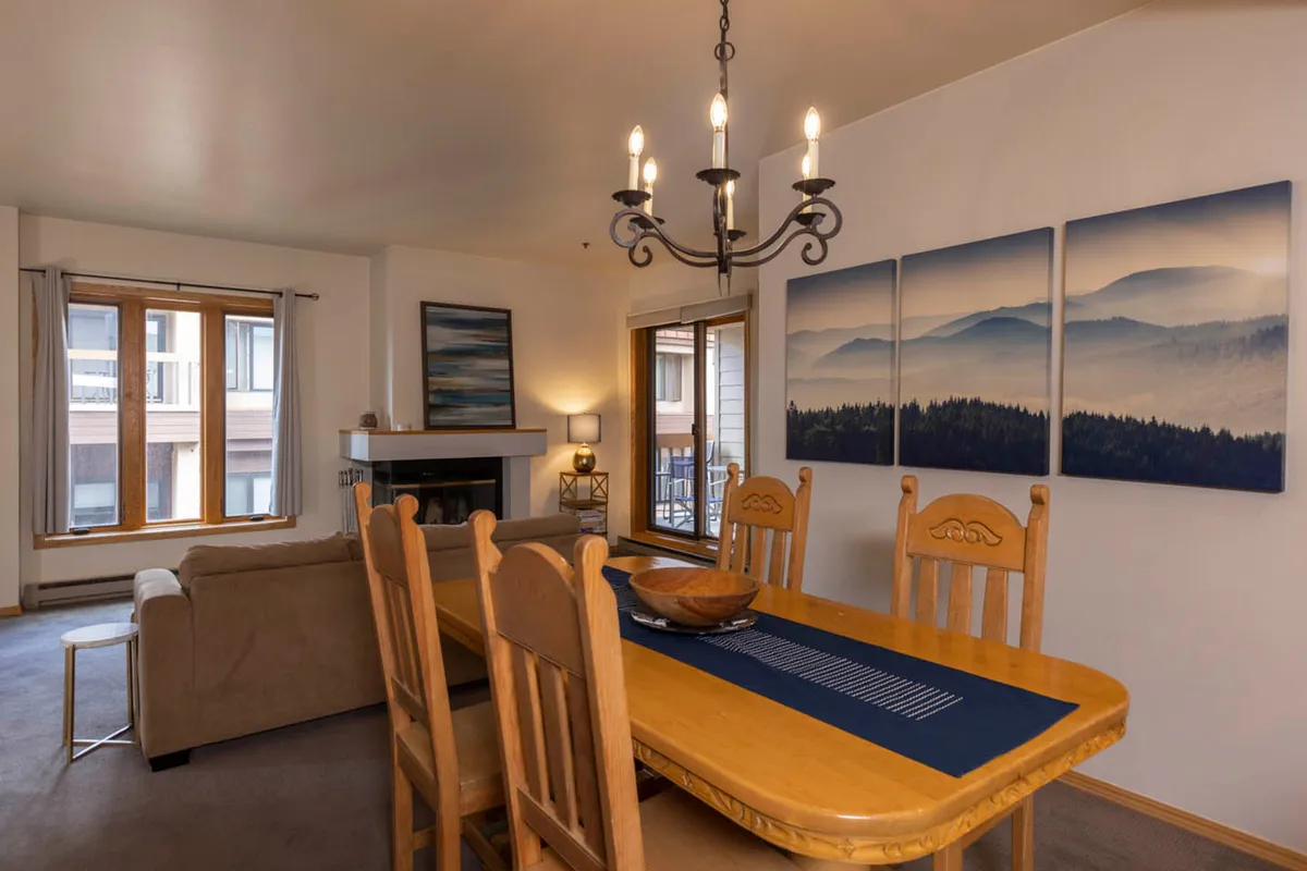A warmly decorated interior with a dining table set near a sofa and fireplace. The space is accented by landscape artwork and a chandelier, evoking a sense of togetherness and comfort ideal for family gatherings and relaxed evenings.