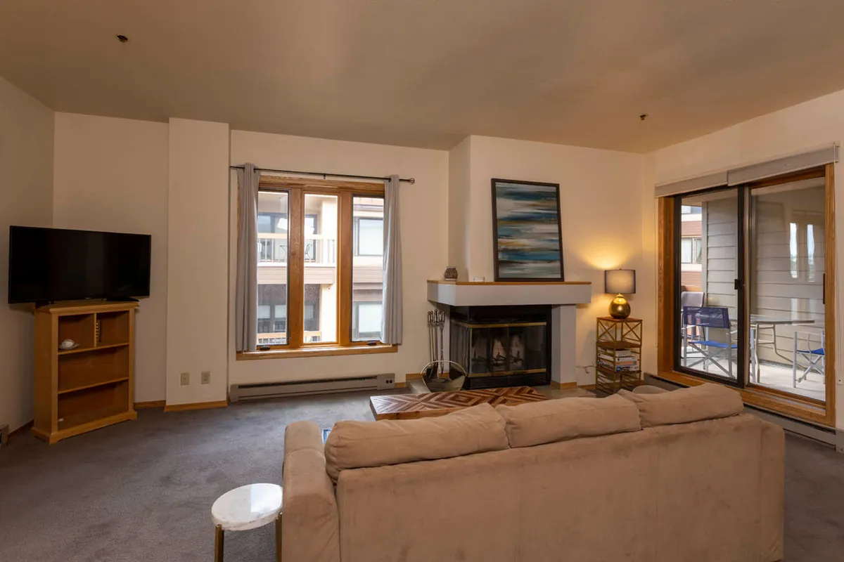 A cozy living room featuring a prominent wood-burning fireplace, a plush sofa positioned near large windows, and a television. The space is warmly lit and arranged for relaxation and socializing, capturing the inviting atmosphere of a mountain retreat.