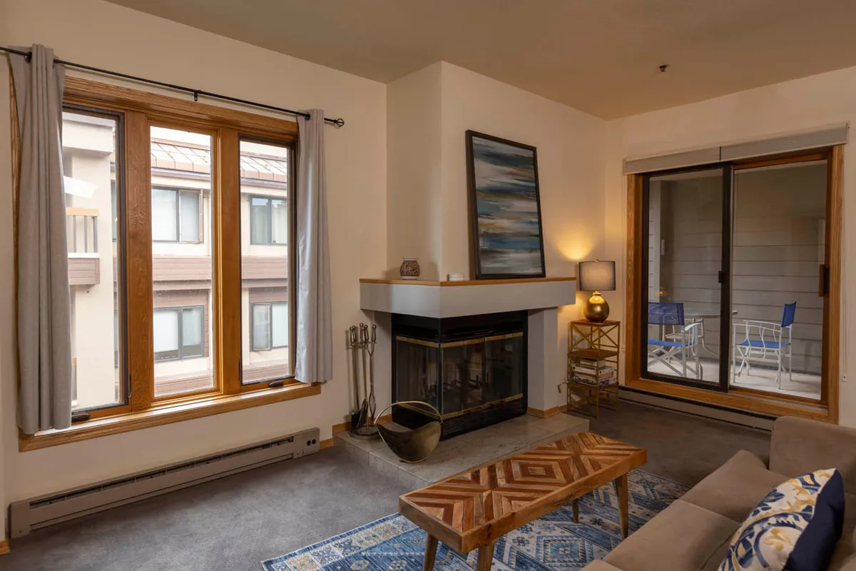 The image showcases a cozy condo living room in Durango, Colorado, featuring a crackling wood-burning fireplace as the focal point. Large windows bathe the space in natural light, highlighting a stylish wooden coffee table, comfortable sofa, and thoughtfully selected artwork that adds a touch of mountain inspiration. The setting invites intimate gatherings, relaxation with a good book or cup of coffee, and a sense of warmth and comfort after a day of outdoor adventure.