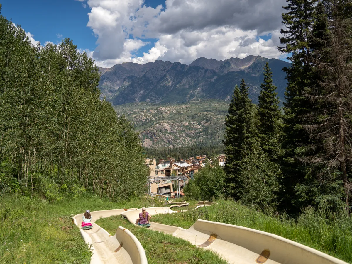 The scene shows people enjoying thrilling rides down a slide with the breathtaking mountain range rising behind them. Lush trees and natural beauty surround the activity, making it clear that outdoor adventure and fun are just steps away.
