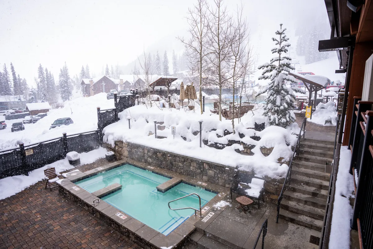 An inviting outdoor hot tub set amidst a snow-covered landscape, with a tree dusted in snow and a staircase leading to the tub. The surrounding area exudes tranquility and warmth, with steam rising from the water as the winter scenery creates a magical atmosphere.