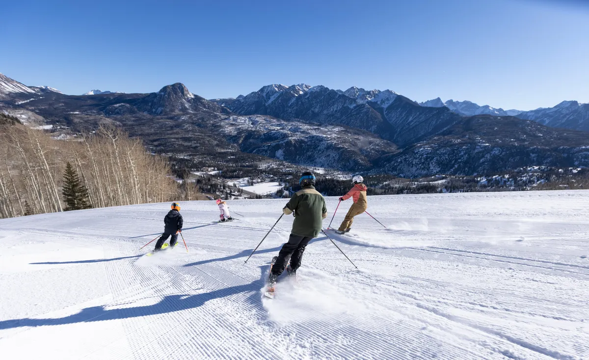 In this dynamic winter scene, skiers traverse pristine snow in front of a majestic mountain range. The landscape is alive with action, showing both individual thrill and group activities, conveying the excitement and variety of experiences available at the resort. The snowy environment and active participants capture the spirit of outdoor adventure central to the section.