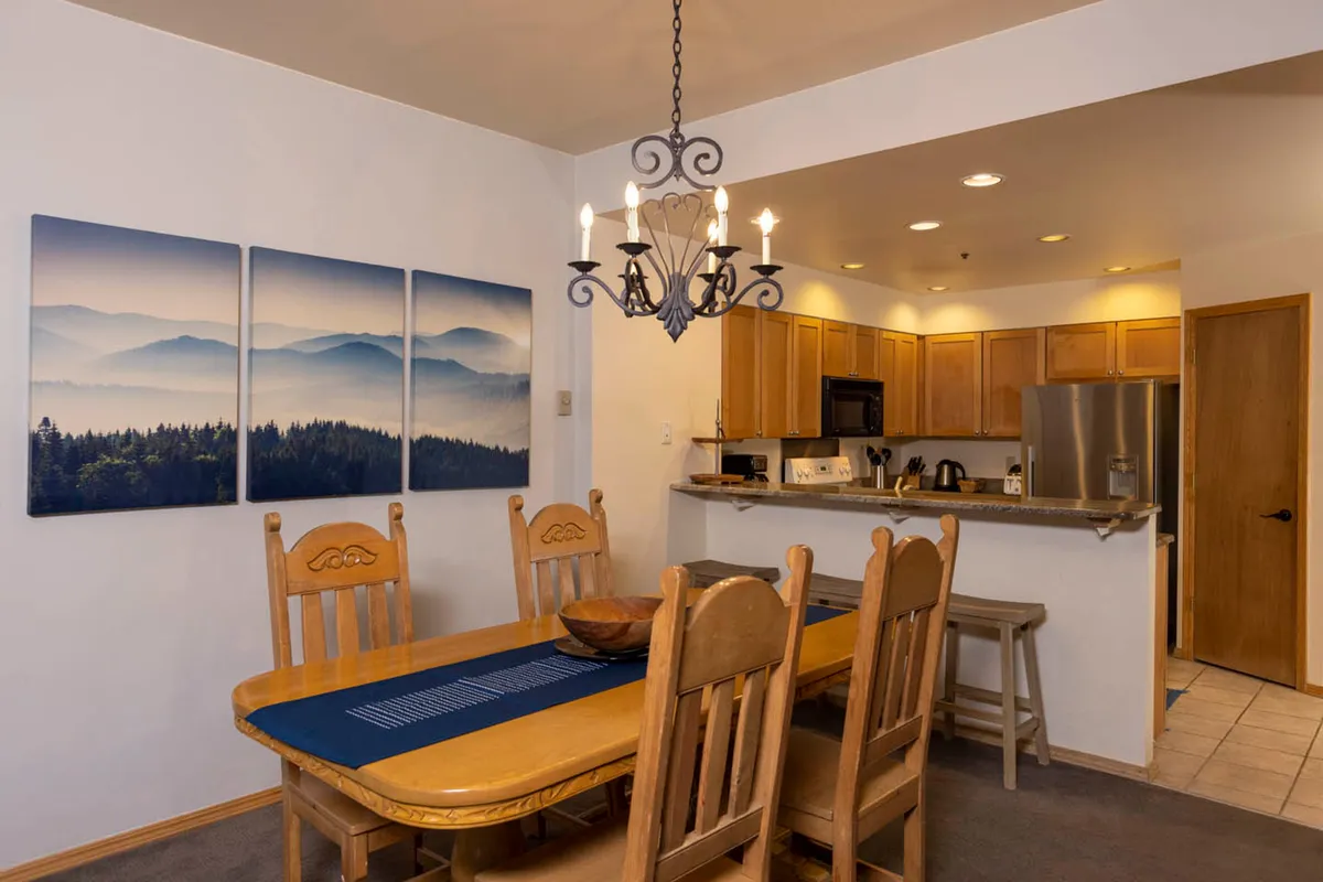The photo features a comfortable dining space with a prominent dining table, elegant chandelier, mountain-themed artwork, and a visible kitchen area. The setting is arranged for group or family dining, emphasizing togetherness.