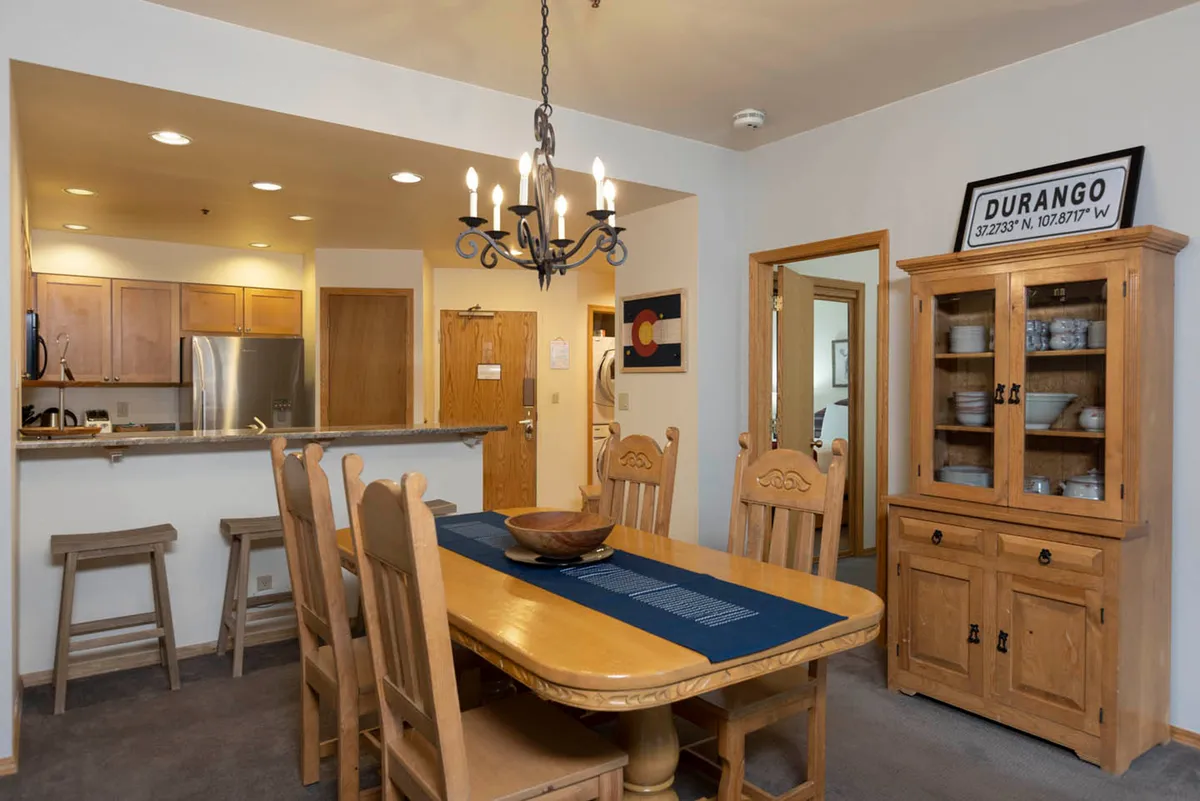 This image captures a spacious dining table set beneath a chandelier, with the kitchen area and a cabinet displaying dishes in the background. The atmosphere is cozy and inviting, perfect for family dinners or group gatherings.