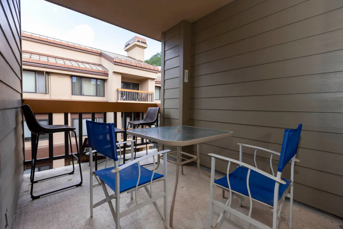 This exterior image features a small balcony with a table and blue chairs, offering a place to enjoy the fresh mountain air and scenic views. The relaxed atmosphere suggests moments of inspiration or breaks between work sessions, with the natural surroundings in view beyond the balcony railing.