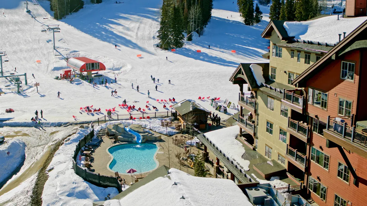 A lively scene featuring an outdoor heated pool, visible ski slopes, a ski lift, and chalet-style buildings. Guests can be seen swimming or relaxing by the pool, while skiers traverse the snowy slopes in the background, all set against a picturesque winter landscape.